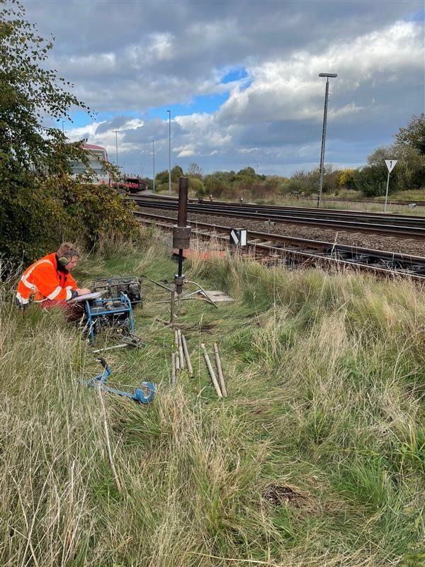 Arbeiter in Warnjacke kniet im hohen Gras neben einem Motorgerät und macht Notizen, dahinter Gestänge und Bahngleise