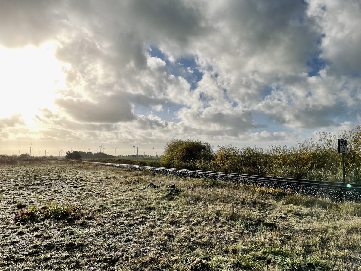 Eingleisige Bahnstrecke durch flaches, frostbedecktes Grasland, Schild mit Kilometerangabe am Gleis, Windräder am Horizont