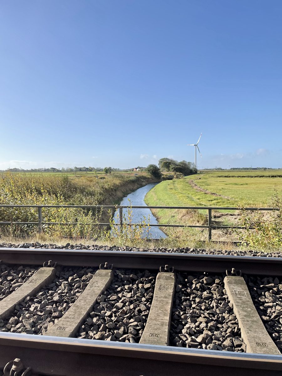 Blick über Schiene und Betonschwellen auf einen wasserführenden Graben mit Geländer, dahinter Grünland und ein Windrad