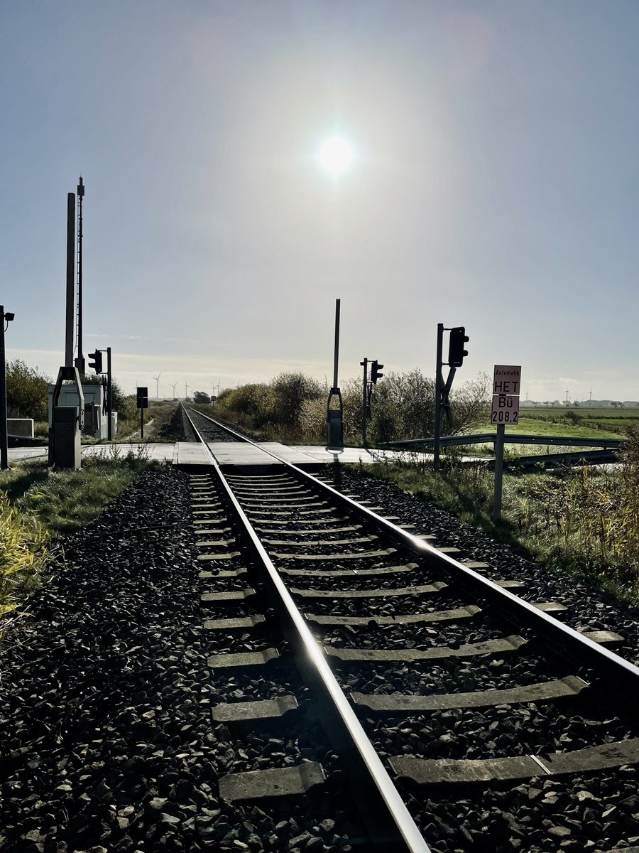 Blick am Gleis entlang auf einen Bahnübergang mit Schranke, Lichtsignalen und Schild Bü 208,2 im Gegenlicht