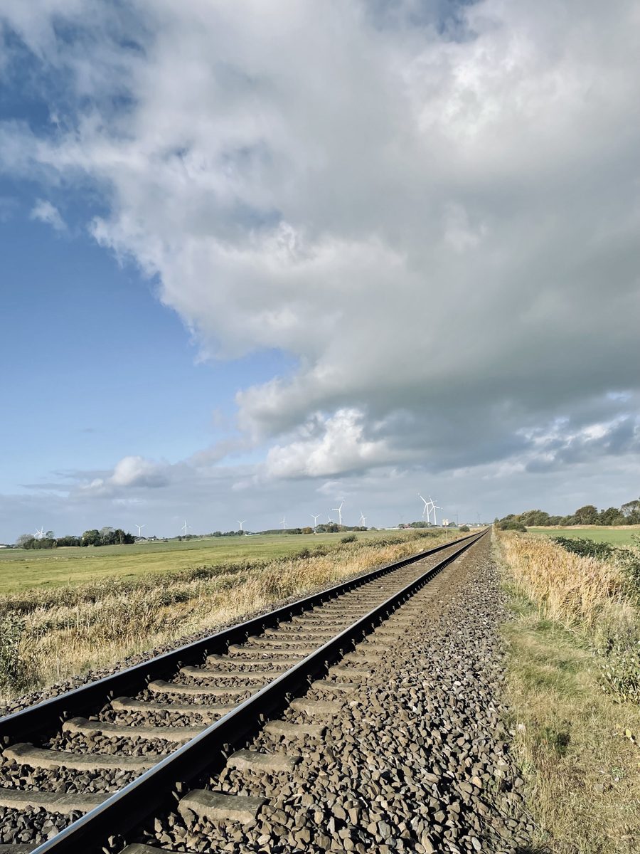 Eingleisige Bahnstrecke im Schotterbett führt gerade durch flache Wiesen, am Horizont eine Reihe Windräder