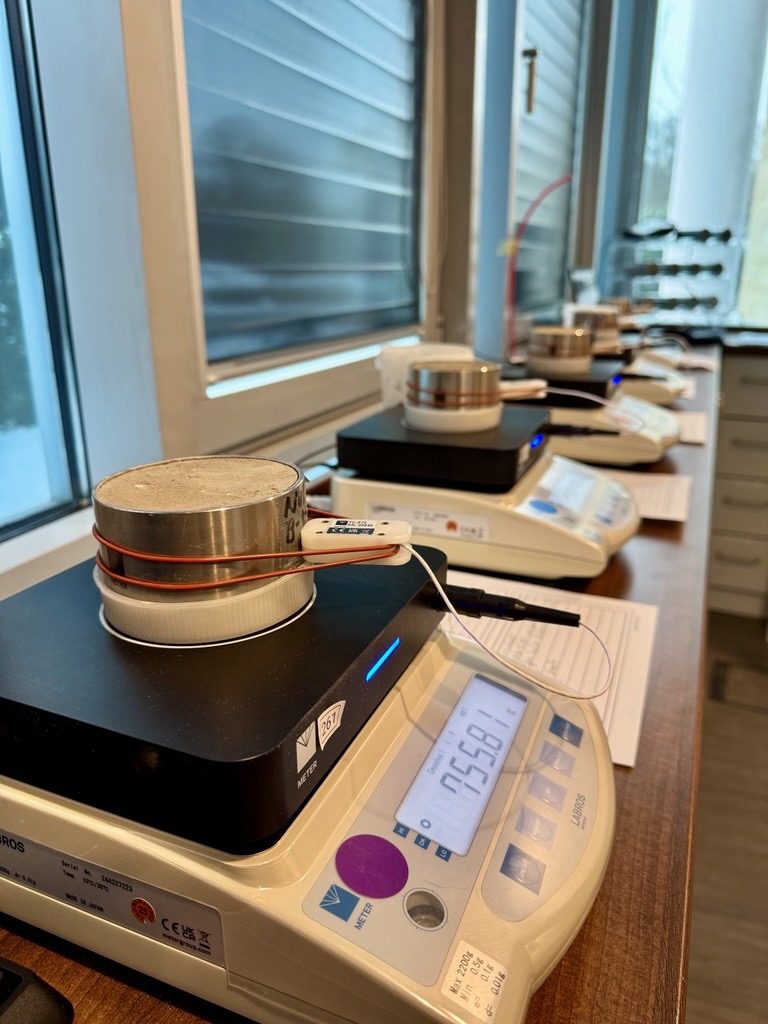 Bodenprobe im Metallzylinder mit aufgesetztem Sensor auf einer Laborwaage, dahinter weitere Proben in Reihe