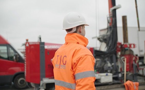 Mitarbeiter von hinten, weißer Helm und orange TIG-Warnjacke, dahinter ein Baugerät mit Mast und ein roter Transporter