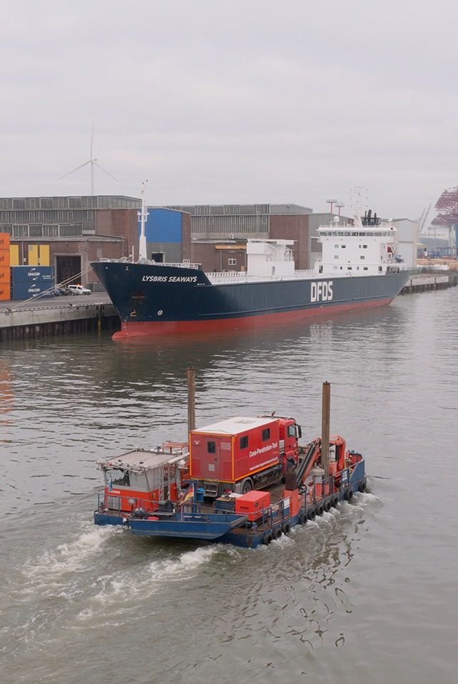 Ponton mit rotem Sondierfahrzeug und zwei aufgestellten Stahlpfählen fährt durch ein Hafenbecken, am Kai ein Frachtschiff